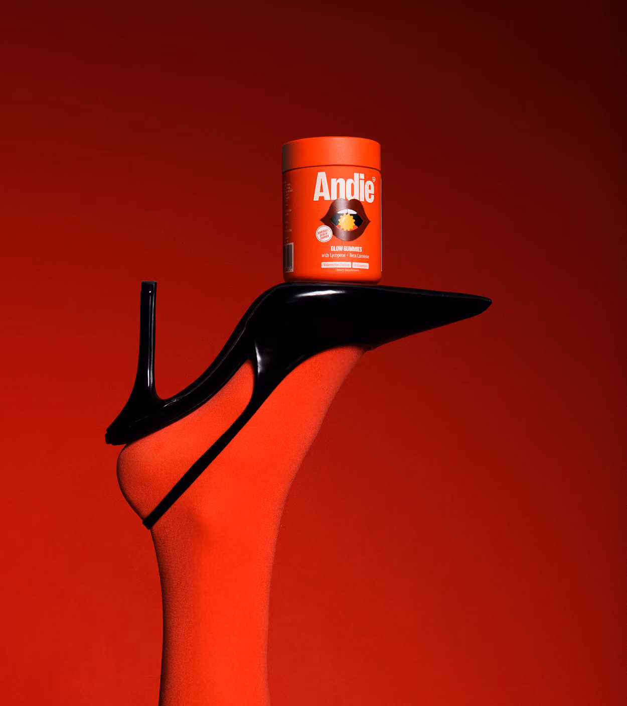 Black high heel shoe worn over a red stockinged foot balancing a red container of Andie Glow Gummies against a red background.