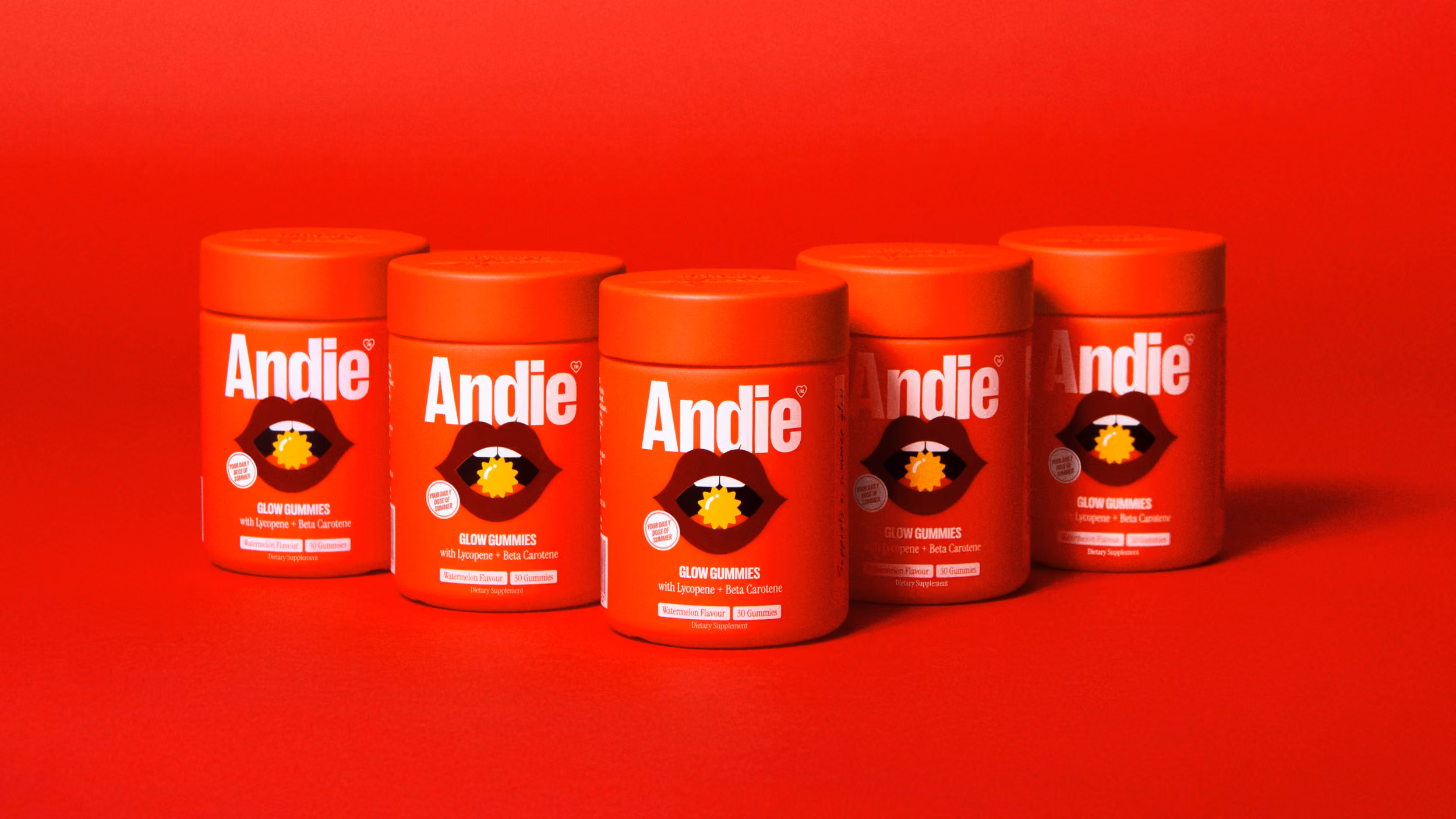 Five red containers of Andie Glow Gummies dietary supplements with watermelon flavor and lycopene plus beta carotene on a red background.