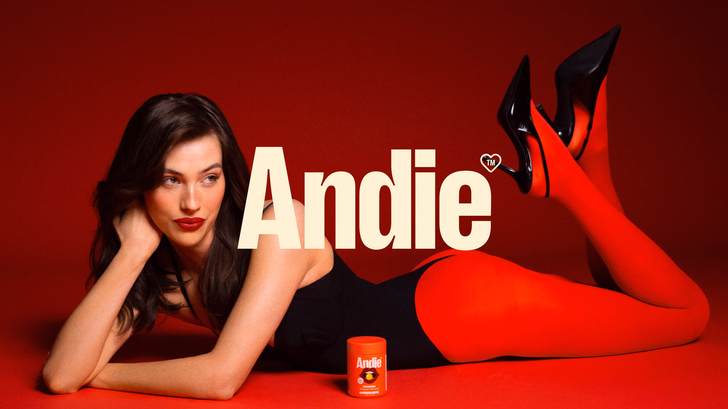 Woman with dark hair wearing red tights and black high heels lying on red floor next to an orange container labeled Andie.