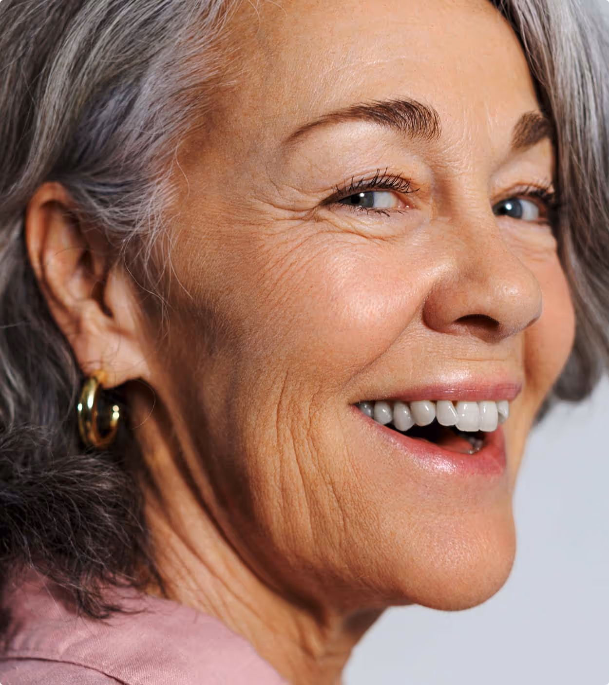 Close-up of a smiling older woman with gray hair, wearing gold hoop earrings and a pink blouse.