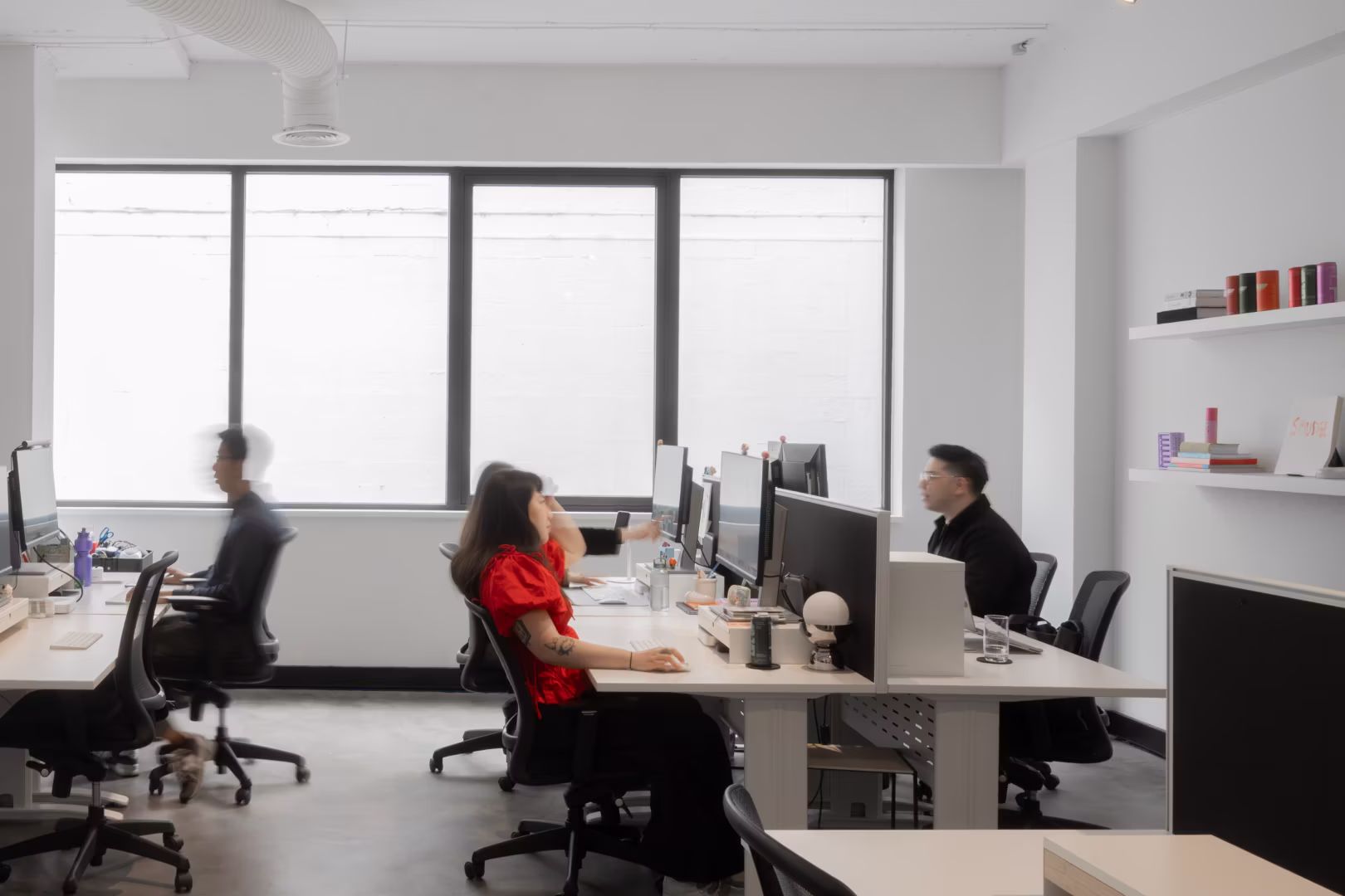Four people working at desks with computers in a bright modern office with large windows.