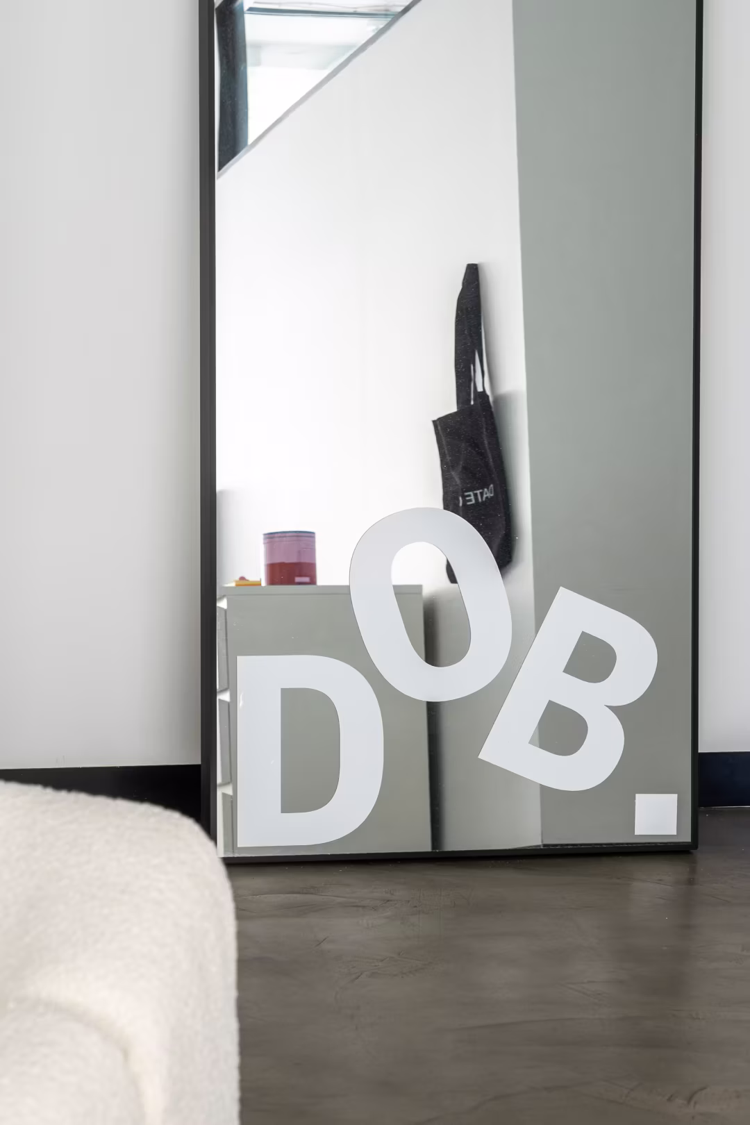 Floor mirror with large white letters DOB. reflecting a cabinet and black bag on wall.