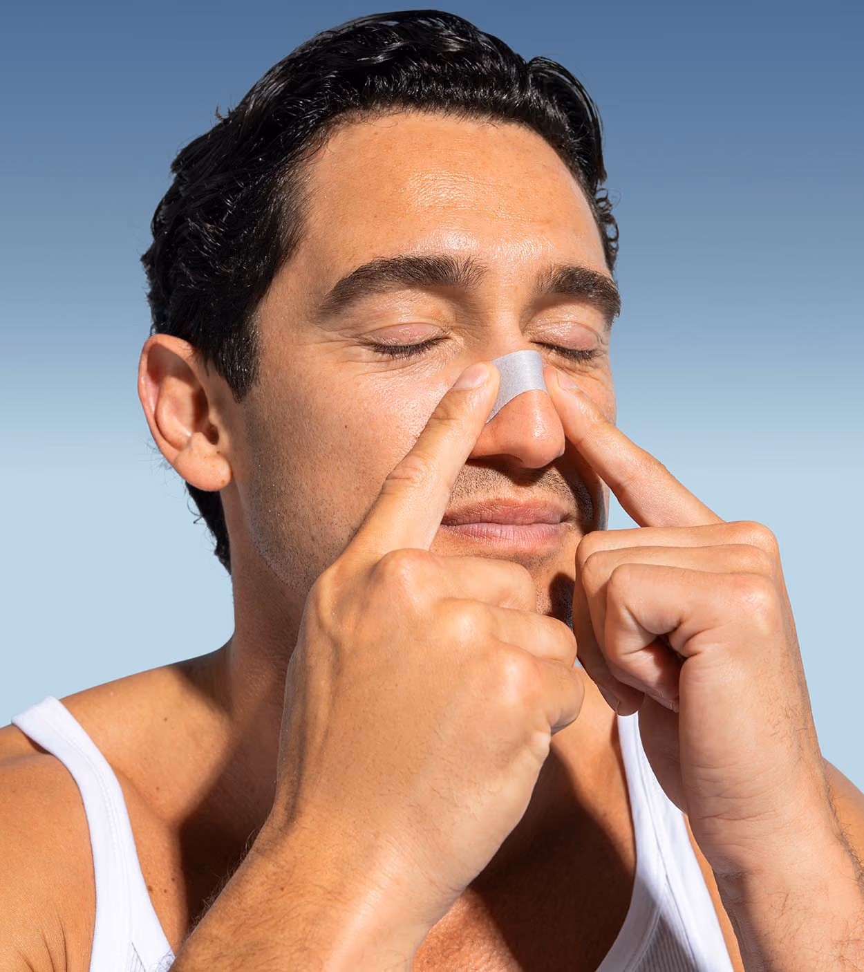 Man with closed eyes placing a nasal strip over the bridge of his nose wearing a white tank top.