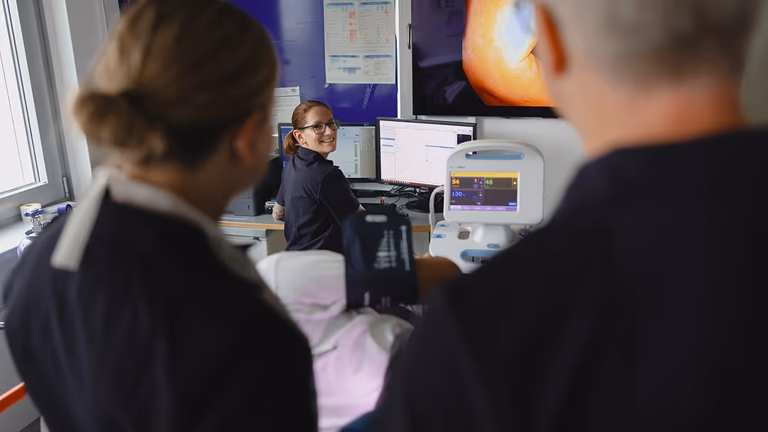 Medizinisches Personal in einem Untersuchungsraum, eine Frau sitzt am Computer und lächelt, während zwei weitere Personen im Vordergrund stehen.