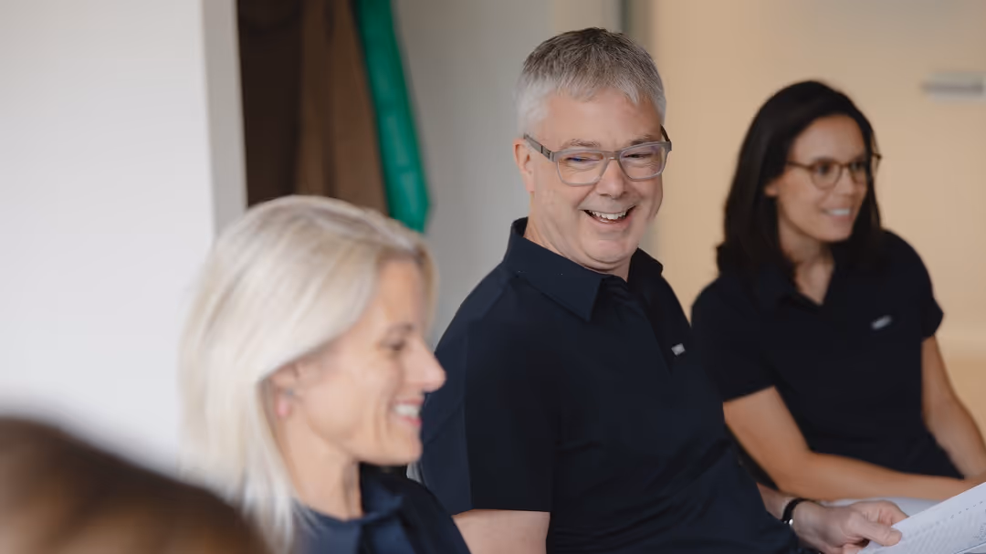 Drei lächelnde Personen sitzen zusammen und wirken entspannt in einer Besprechung.