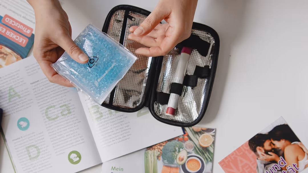 Eine Person hält einen blauen Kühlakku über einem geöffneten medizinischen Notfall-Set mit einem Insulinstift, umgeben von geöffneten Broschüren und Magazinen auf weißem Tisch.