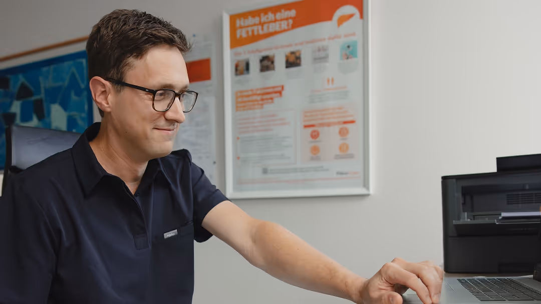 Mann mit Brille arbeitet an einem Laptop an einem Schreibtisch mit einem Informationsposter im Hintergrund.