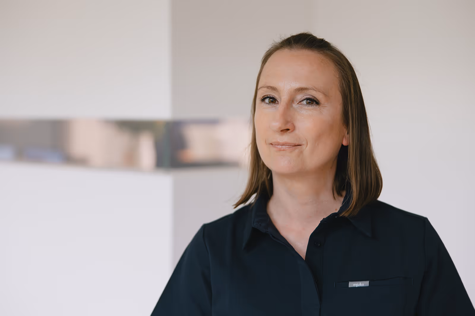 Porträt einer Frau mit mittellangen braunen Haaren, die in einem schwarzen Hemd vor heller Wand steht.
