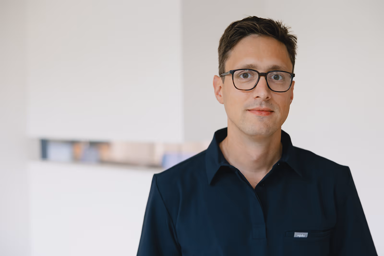 Porträt eines jungen Mannes mit Brille und dunklem Hemd vor unscharfem weißem Hintergrund.