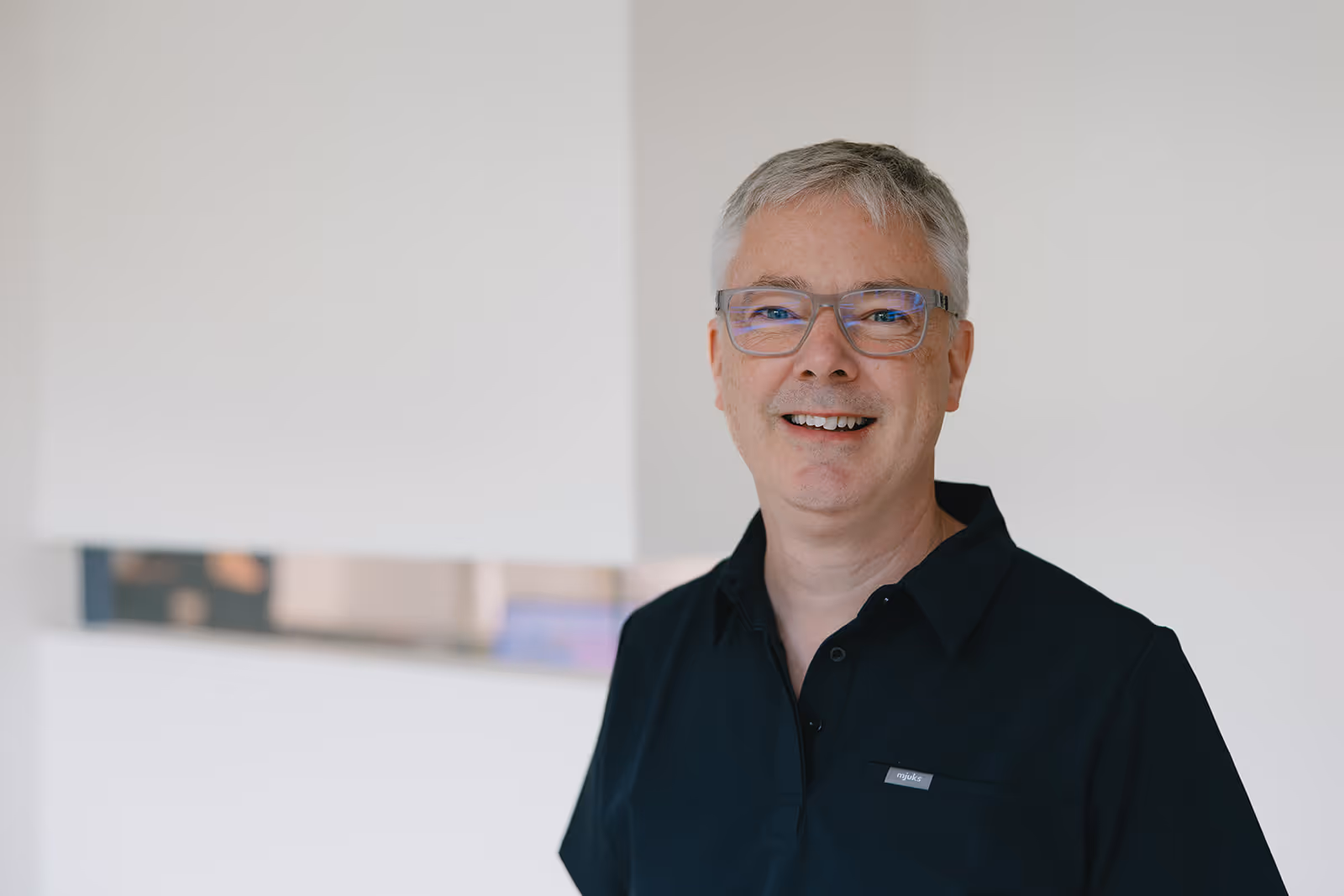 Lächelnder, älterer Mann mit grauem Haar und Brille vor hellem Hintergrund.