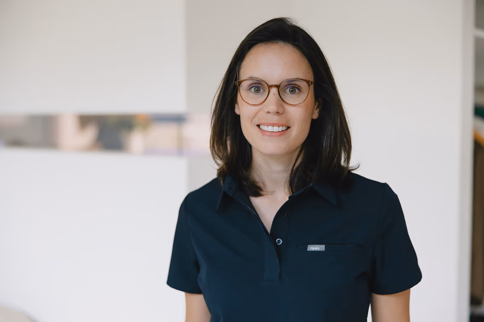 Frau mit dunklen Haaren, die eine Brille trägt und ein schwarzes Hemd vor hellem Hintergrund lächelnd schaut.