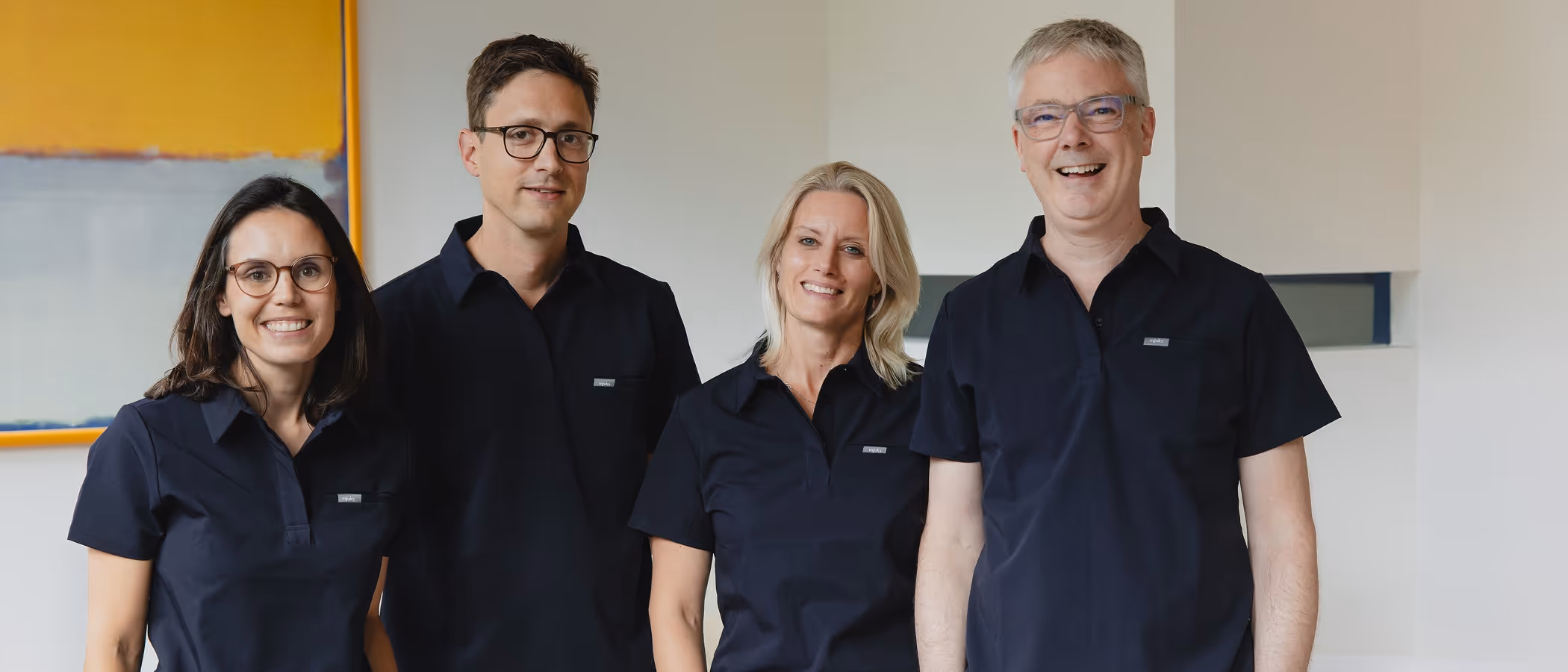 Vier lächelnde Erwachsene, zwei Frauen und zwei Männer, alle in dunkelblauen Poloshirts stehend vor weißer Wand mit abstraktem Bild.