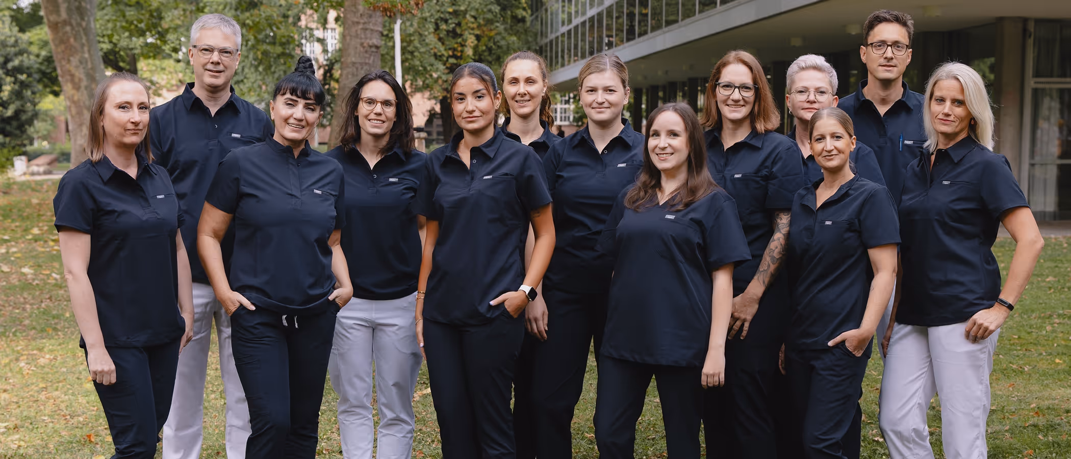 Gruppenfoto von elf Frauen und zwei Männern in medizinischer Arbeitskleidung im Freien vor einem Gebäude.