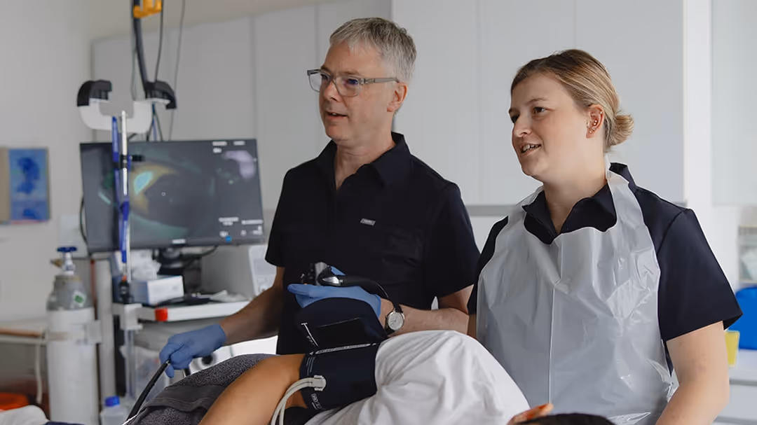 Zwei medizinische Fachkräfte im Gespräch während einer Untersuchung eines liegenden Patienten mit medizinischer Ausrüstung im Hintergrund.