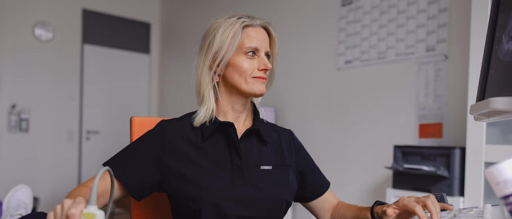 Eine blonde Frau in schwarzer Arbeitskleidung bedient ein medizinisches Gerät in einem Behandlungszimmer.