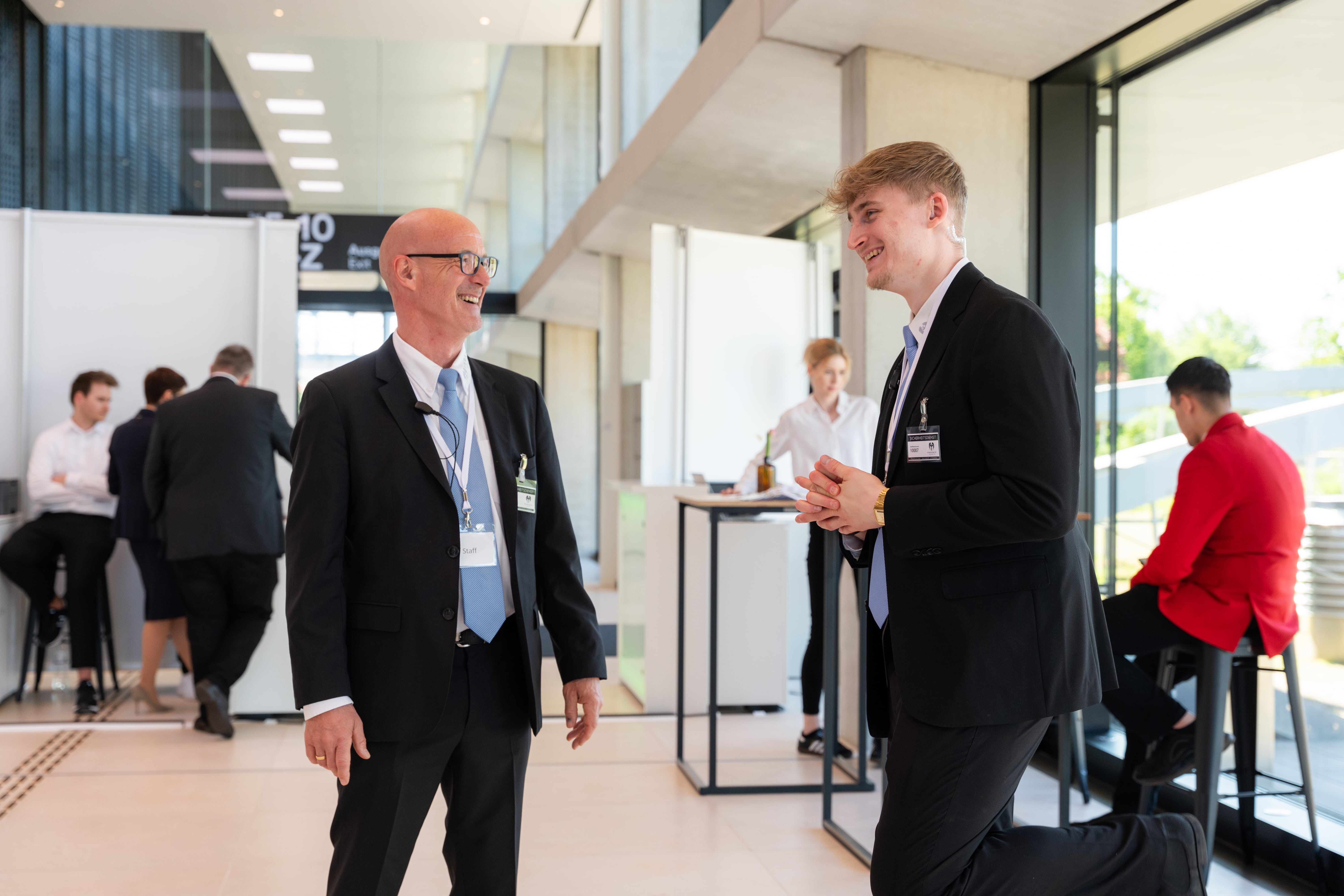 Two men in suits smiling and conversing inside a modern office with other people in the background.