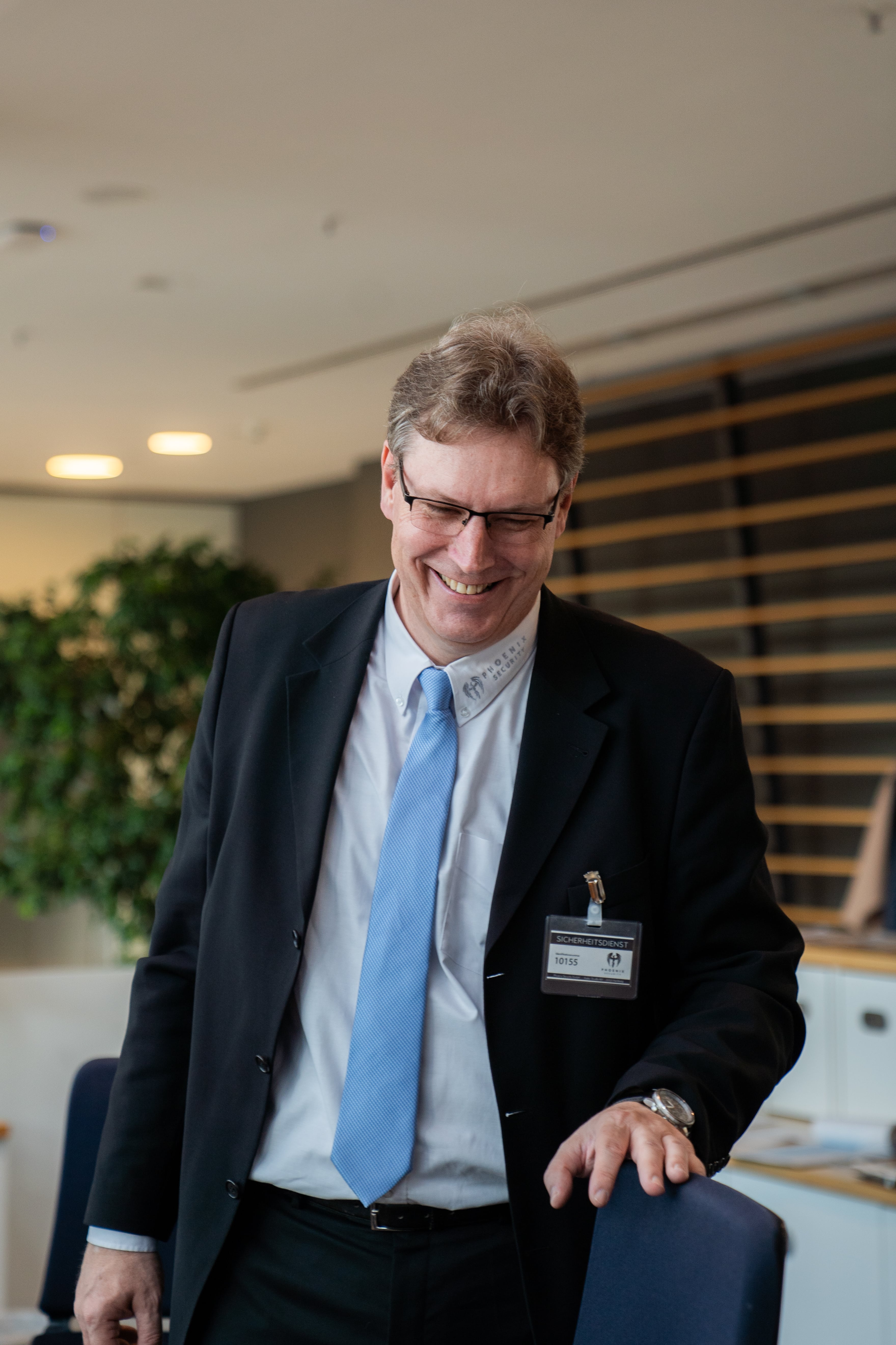 Smiling man in glasses wearing a black suit and blue tie standing in an office, with a security badge clipped to his jacket.