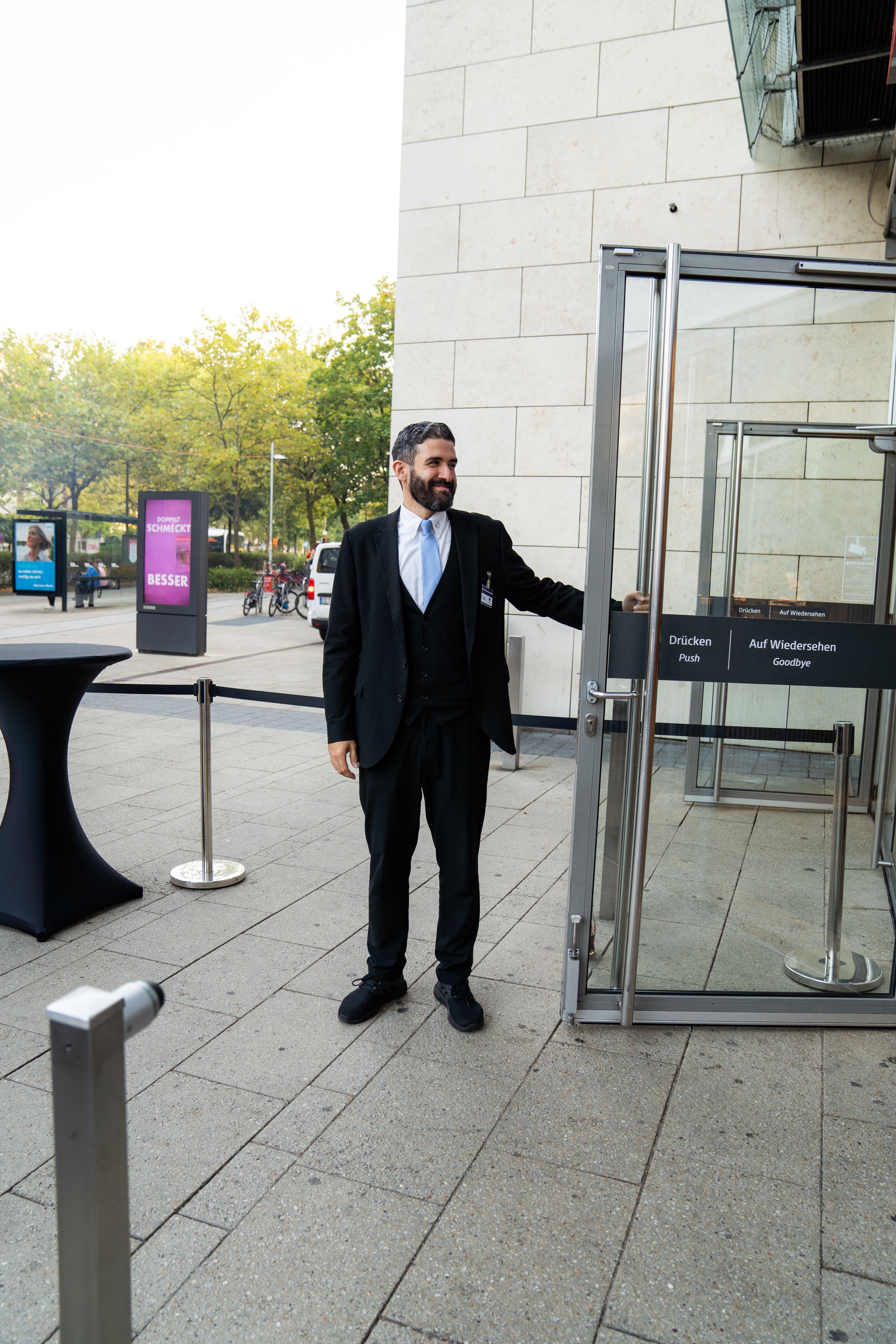 Man in a black suit holding open a glass door outside a building.