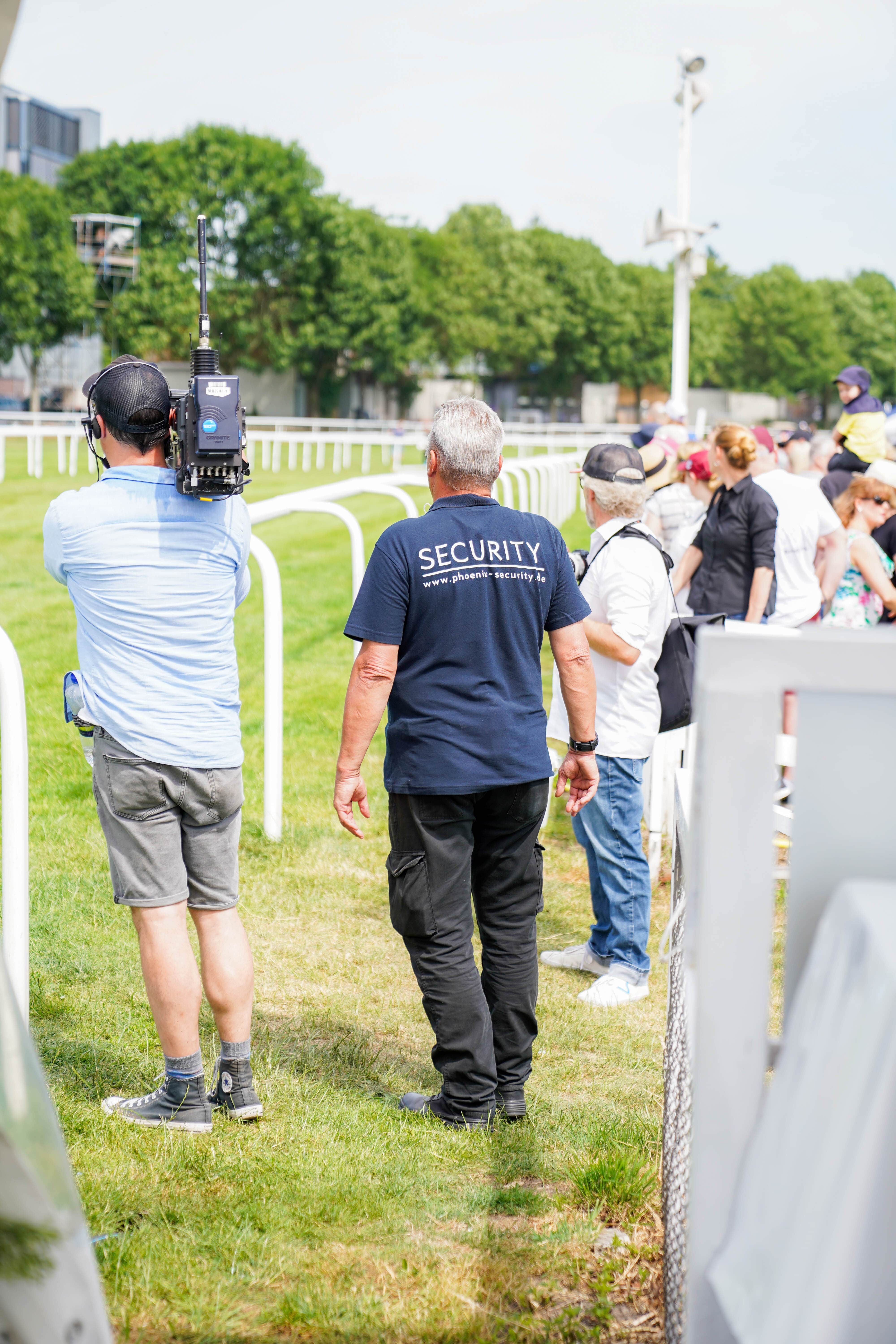 Man wearing a navy blue security shirt and another man filming with a handheld camera at an outdoor event with a crowd in the background.