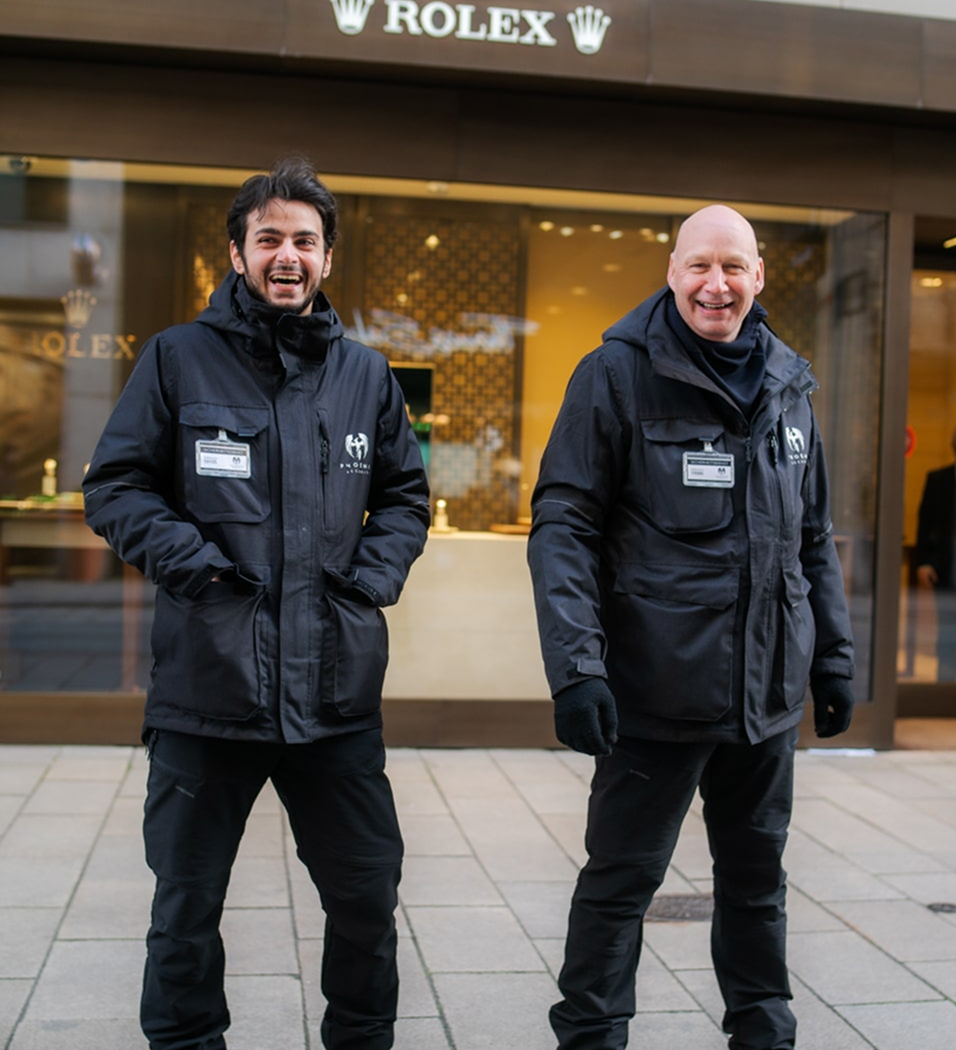 Two smiling men wearing black jackets and security badges standing outside a Rolex store.