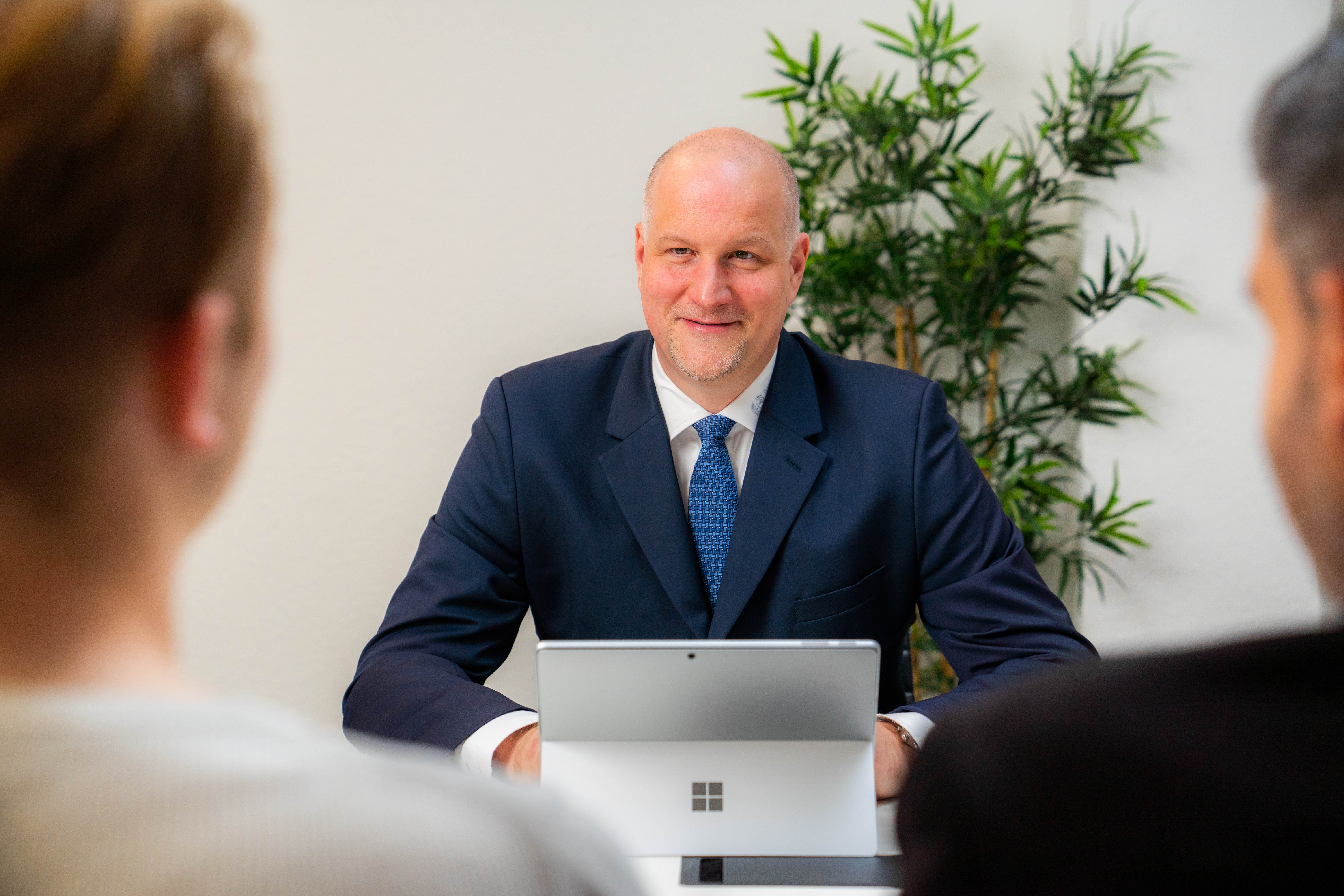 Bald man in a navy suit and blue tie sitting at a table with a Microsoft Surface device, smiling and talking to two people opposite him.