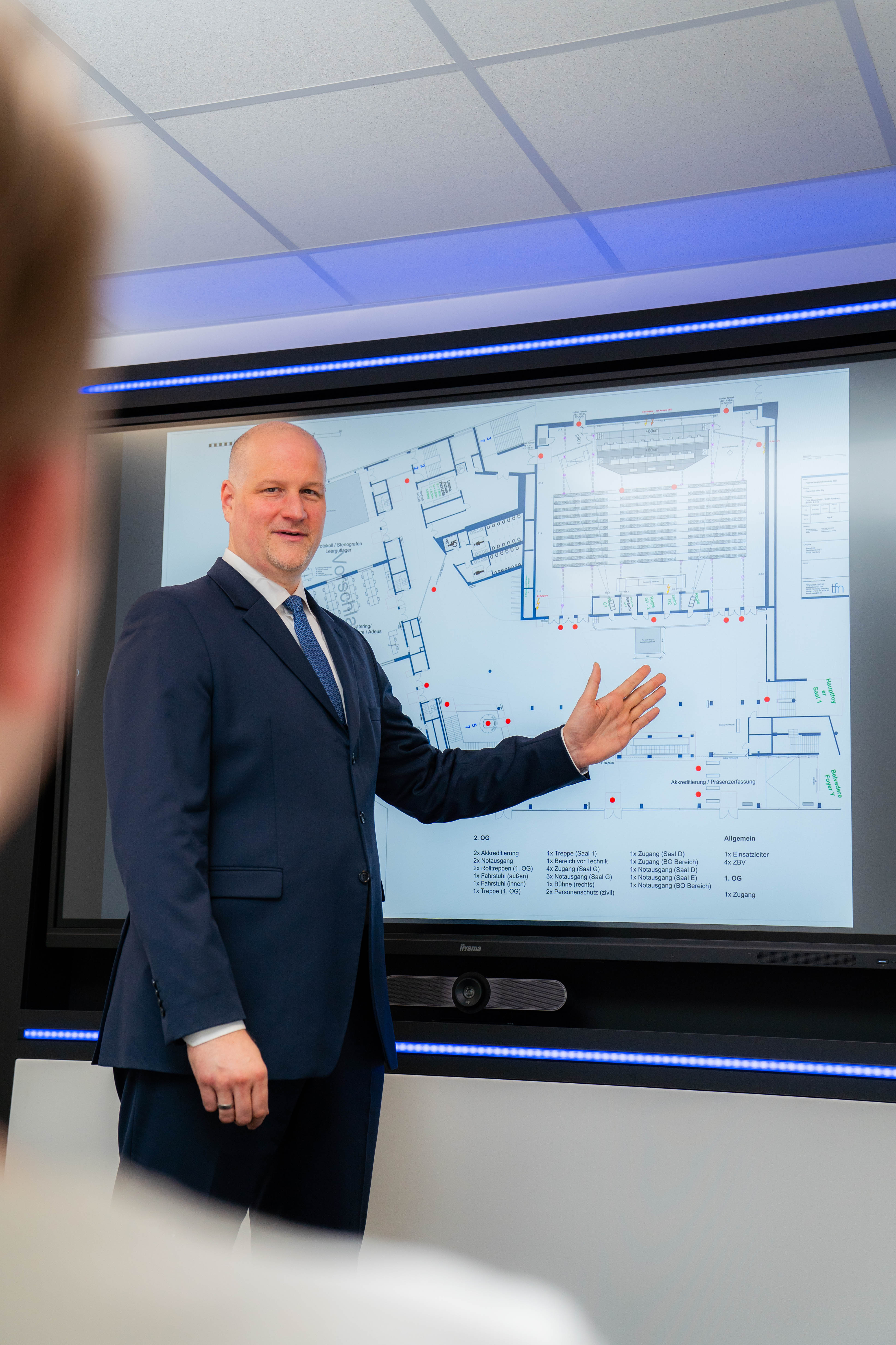 Man in navy suit presenting an architectural floor plan on a large screen in a meeting room.