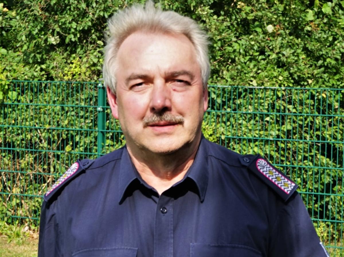 Middle-aged man with light hair and mustache wearing a dark uniform shirt standing outdoors in front of a green fence and foliage.