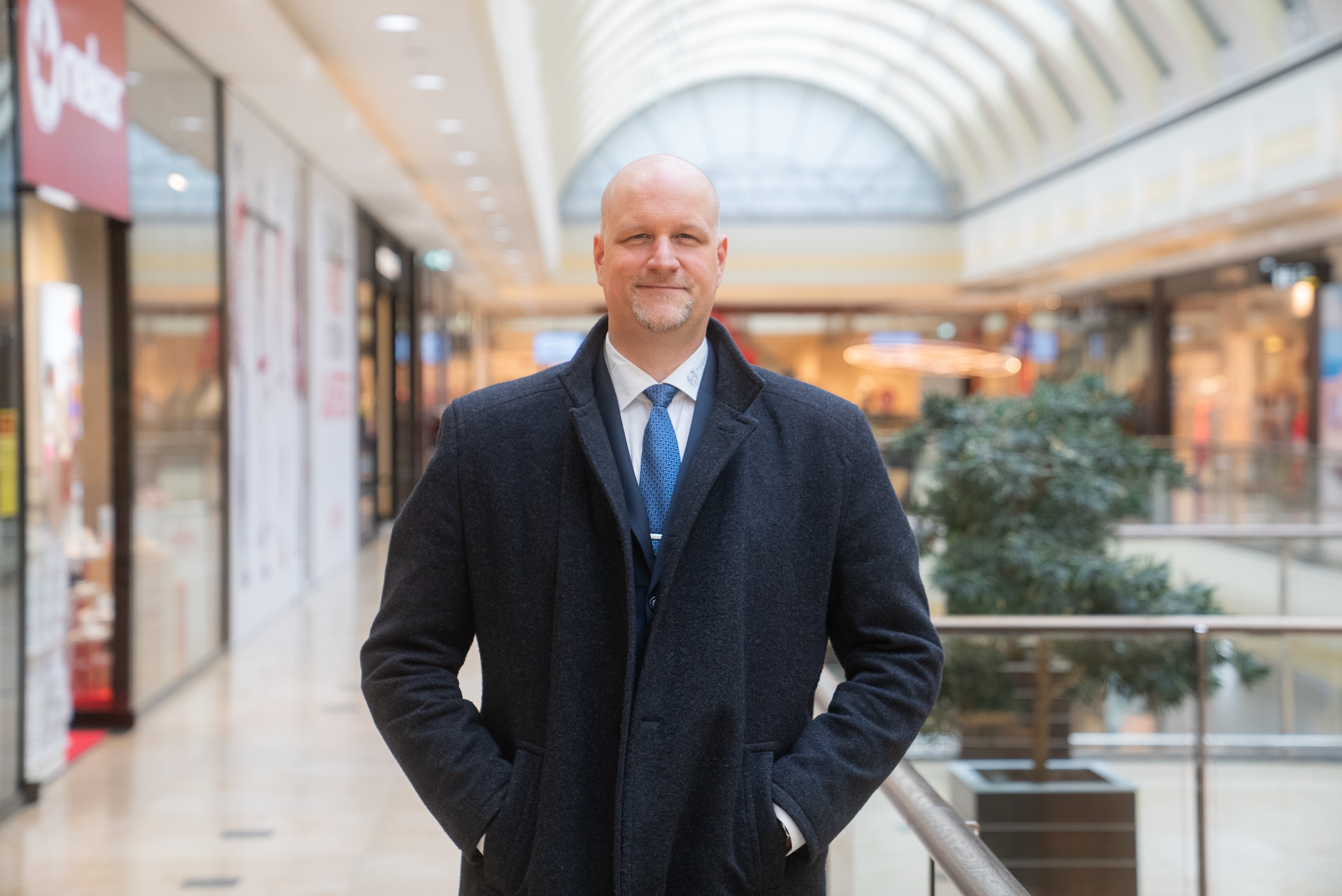 Smiling bald man in a dark coat and blue tie standing indoors in a modern shopping mall.