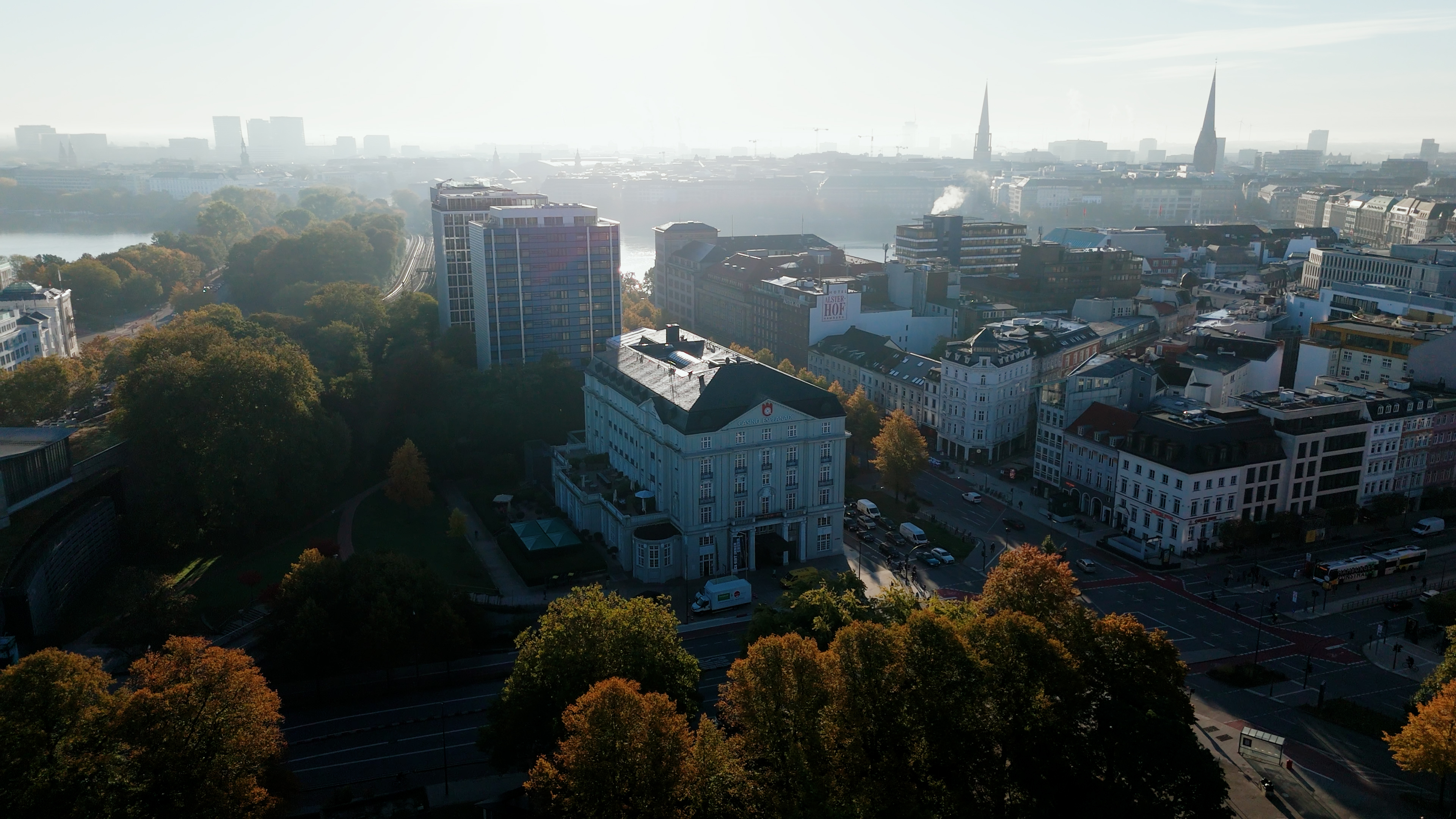 Luftaufnahme einer Stadt mit herbstlich gefärbten Bäumen, mehreren Gebäuden und einer nebligen Skyline im Hintergrund.