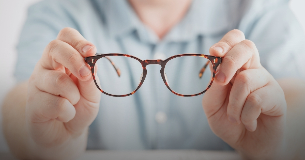 Eine Person hält eine braun getigerte Brille mit beiden Händen vor einem verschwommenen Hintergrund.