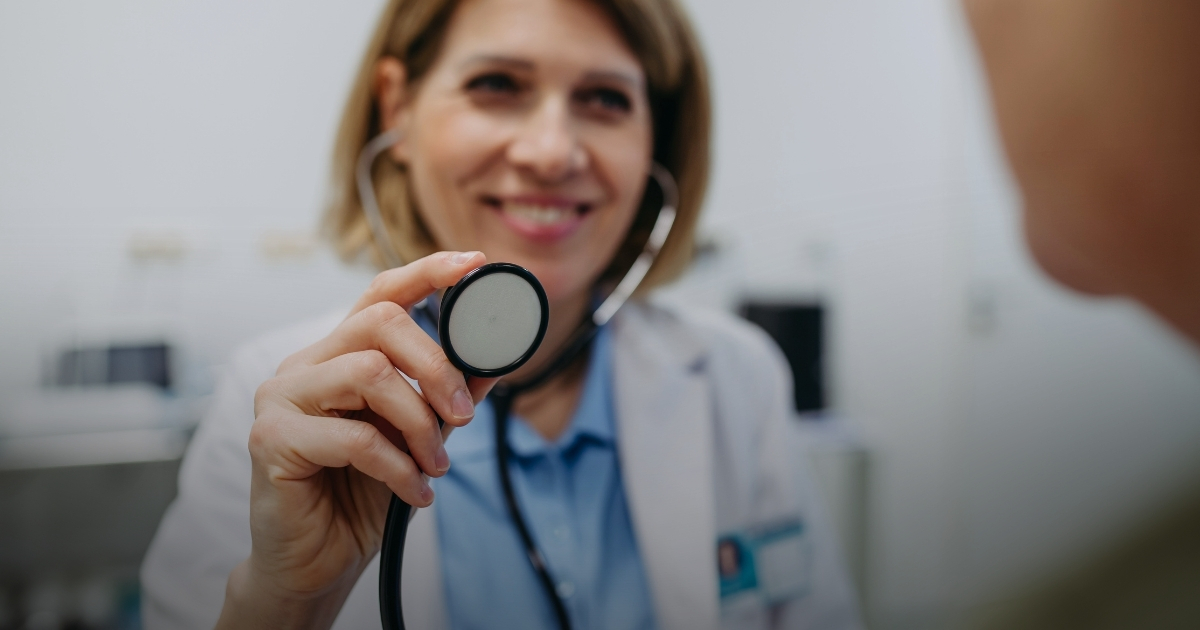 Ärztin hält Stethoskop in den Vordergrund, lächelt und trägt weißen Kittel.