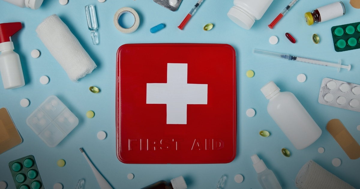 Rotes Erste-Hilfe-Set mit weißem Kreuz umgeben von Medikamenten, Verbänden, Spritzen und Pillen auf blauem Hintergrund.