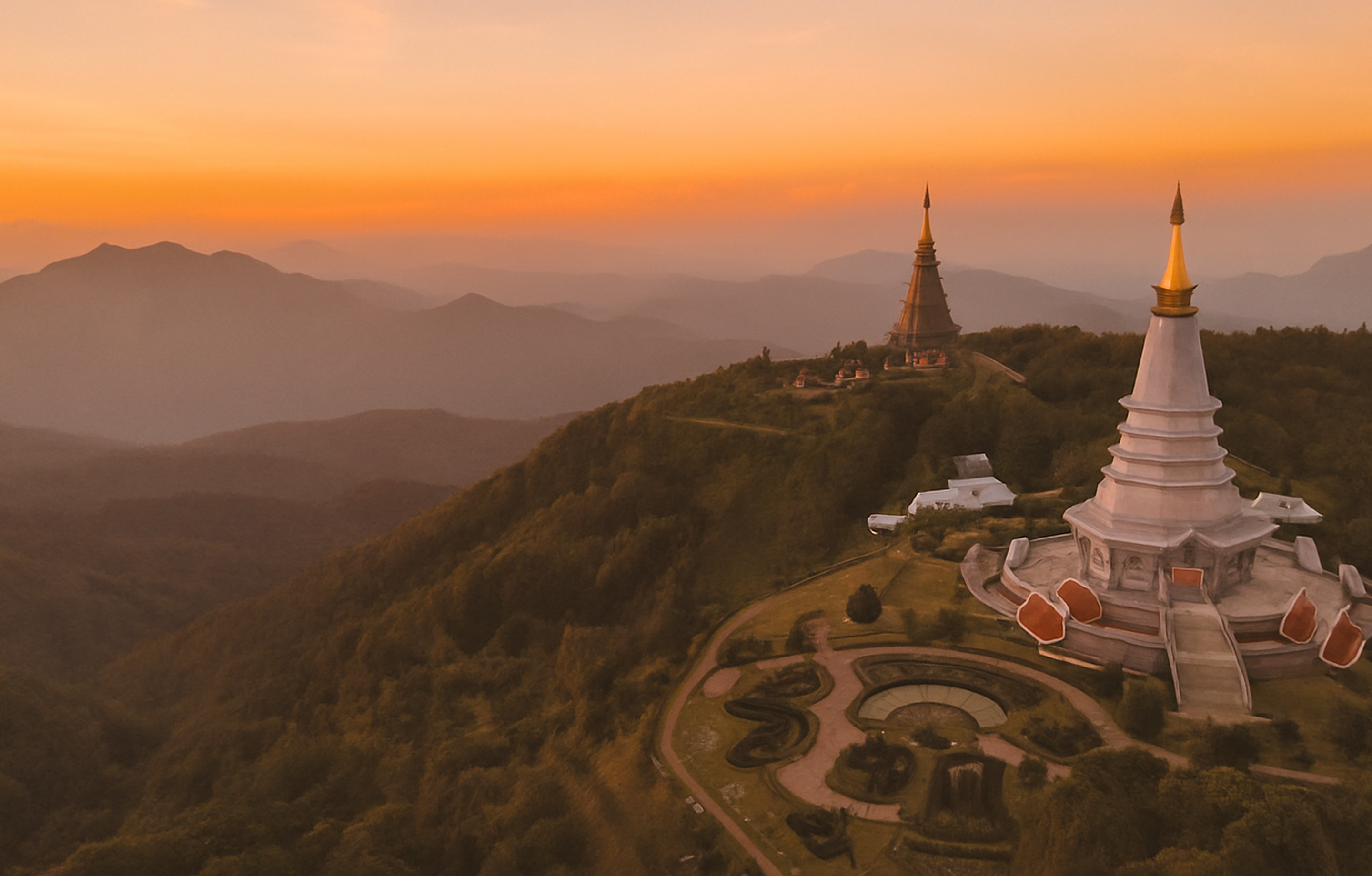 Sunset over mountain landscape with two large Buddhist stupas surrounded by greenery.