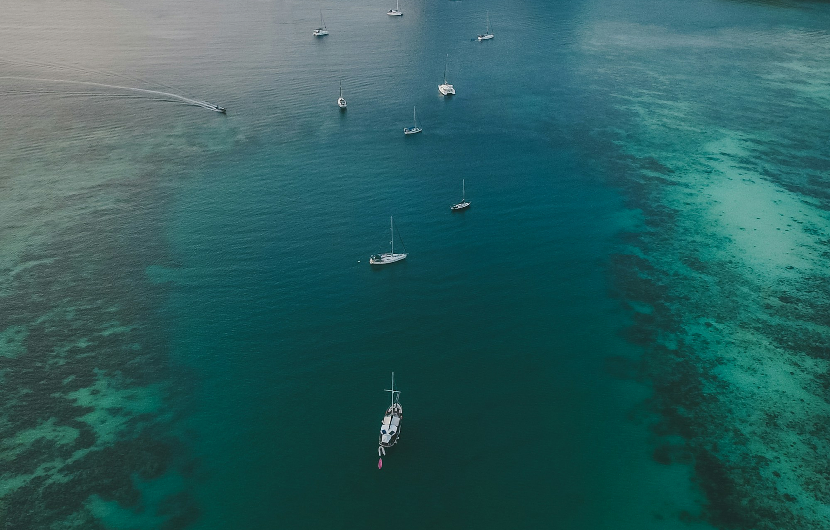 Aerial view of several sailboats anchored in clear blue-green ocean water with visible coral reefs.