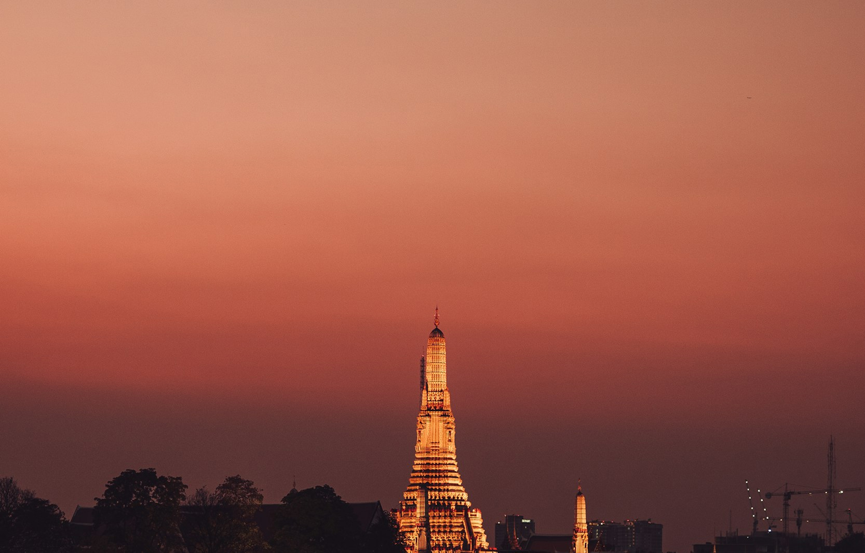 Illuminated temple spire against a warm orange-red sunset sky with silhouettes of trees and city buildings.