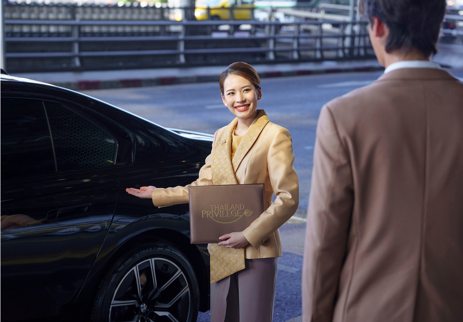 A smiling woman in a beige uniform holding a Thailand Privilege folder gestures invitingly toward a black car while a man in a brown suit stands nearby.