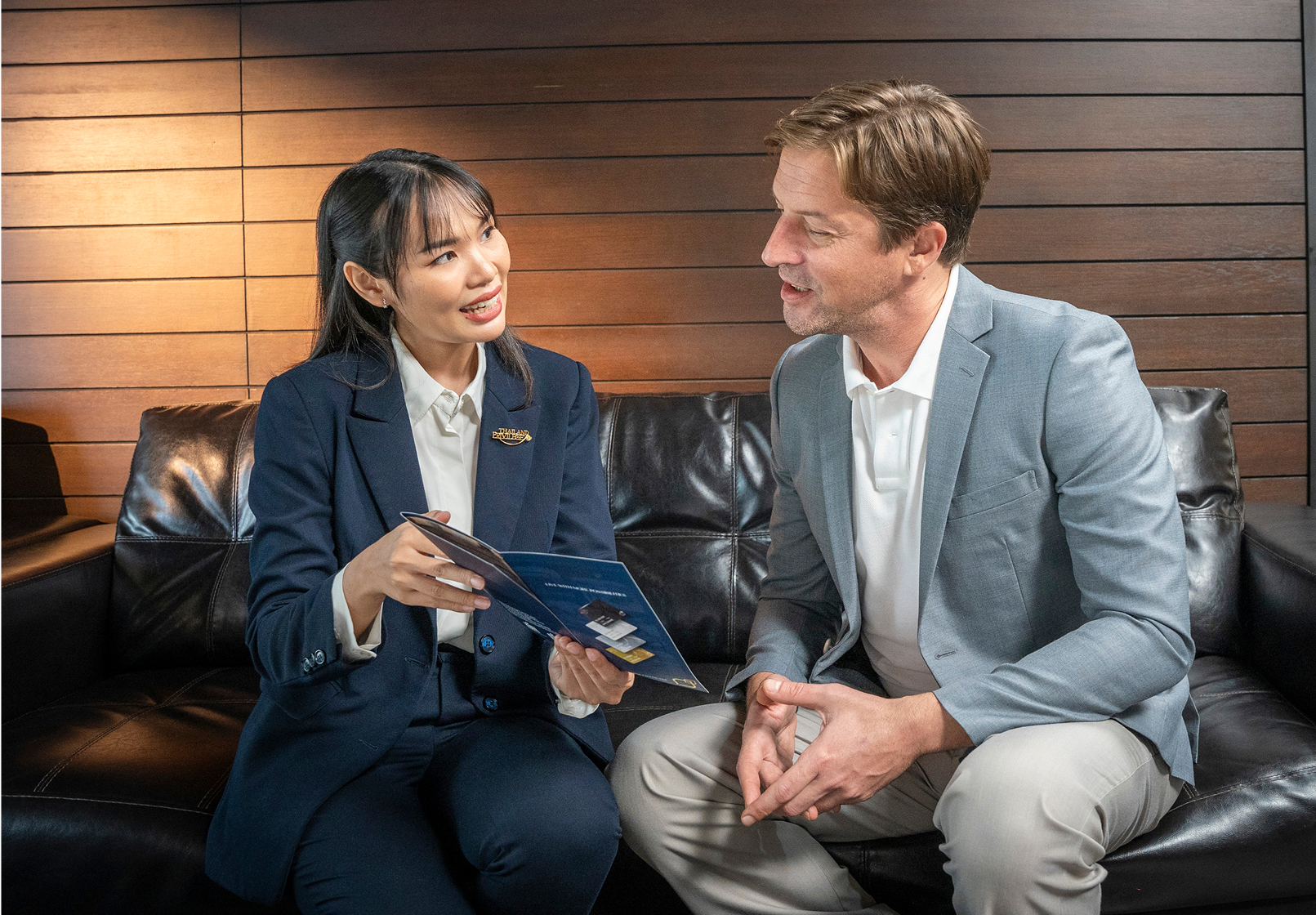 A woman in a navy suit shows a brochure to a man in a light gray blazer while sitting on a black leather couch.