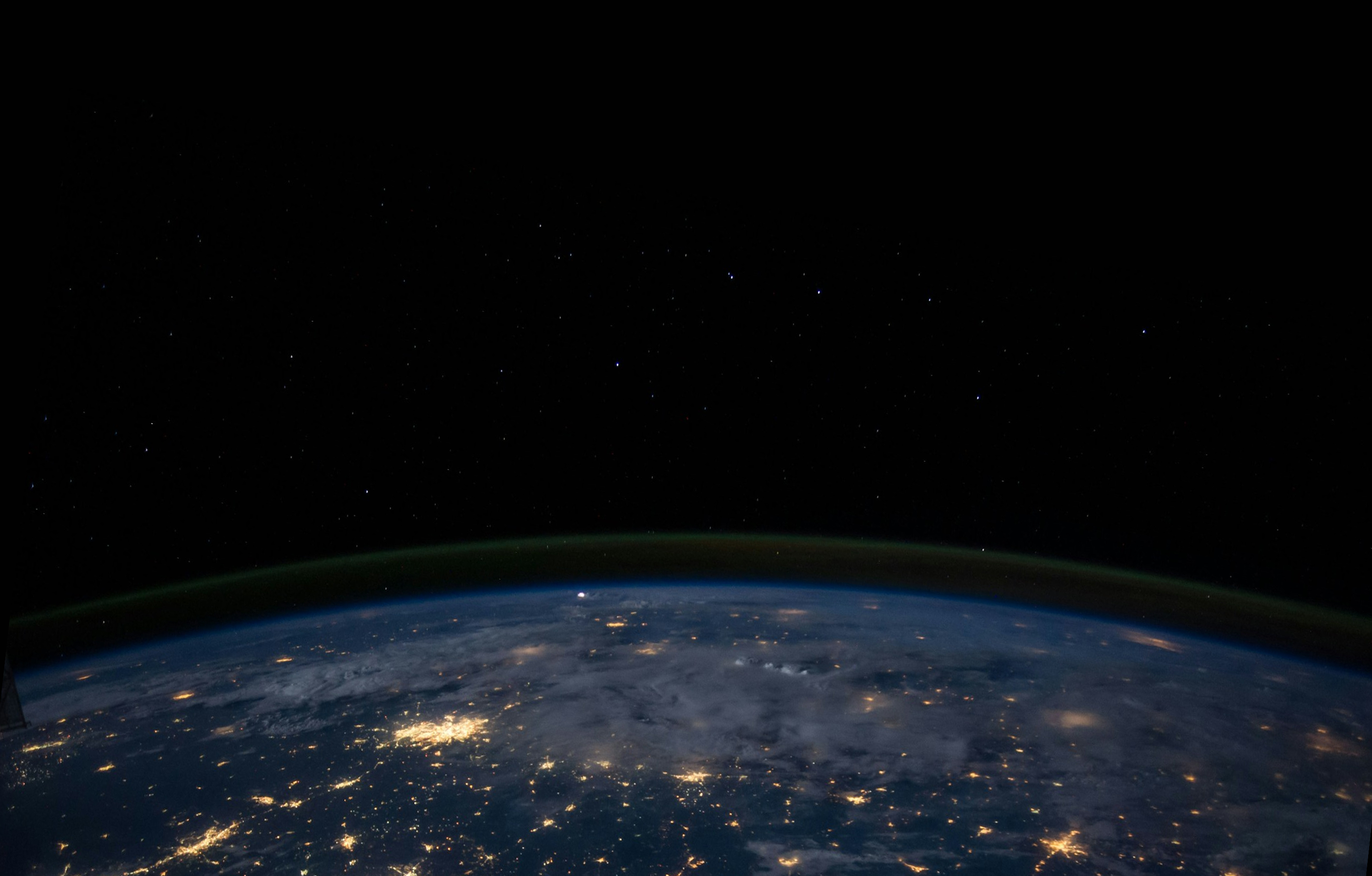 View of Earth from space at night showing city lights and a faint green atmospheric glow under a starry sky.