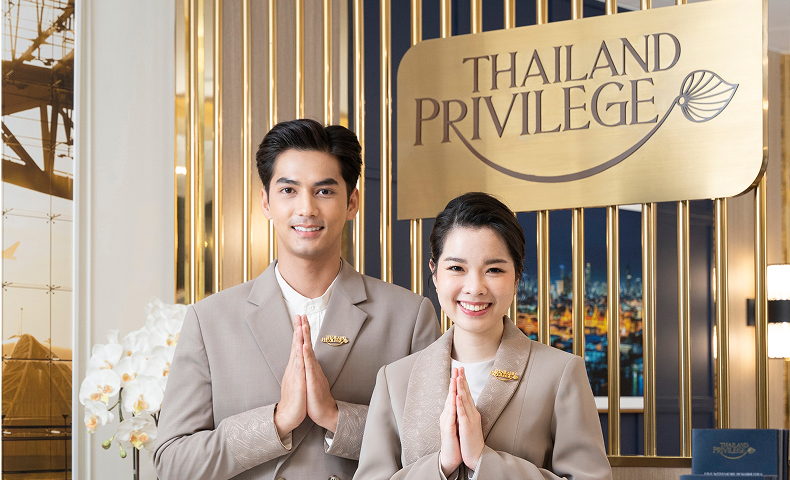 Smiling man and woman dressed in beige uniforms performing the traditional Thai greeting with palms together in front of a gold sign reading Thailand Privilege.