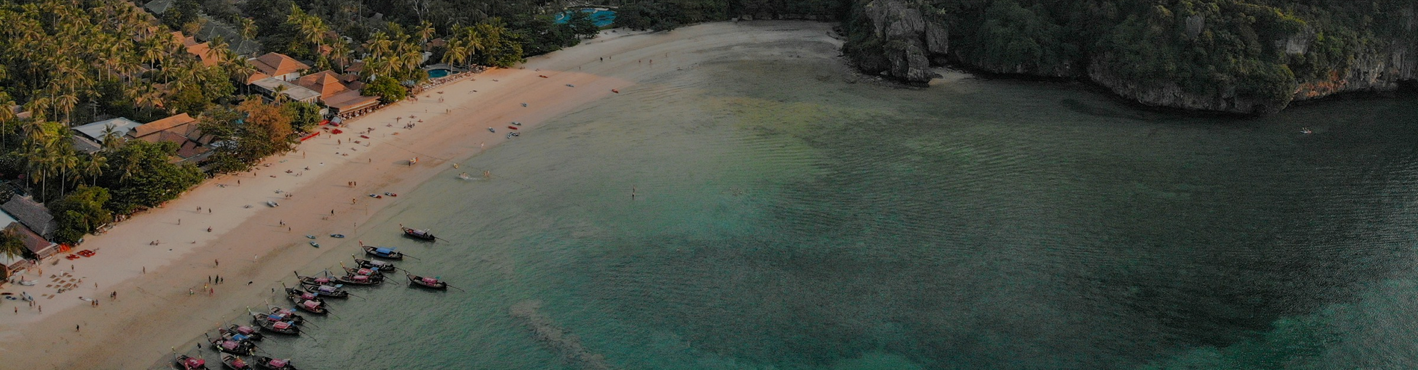 Aerial view of a tropical beach with beachfront buildings, palm trees, boats docked near the shore, and clear shallow water.