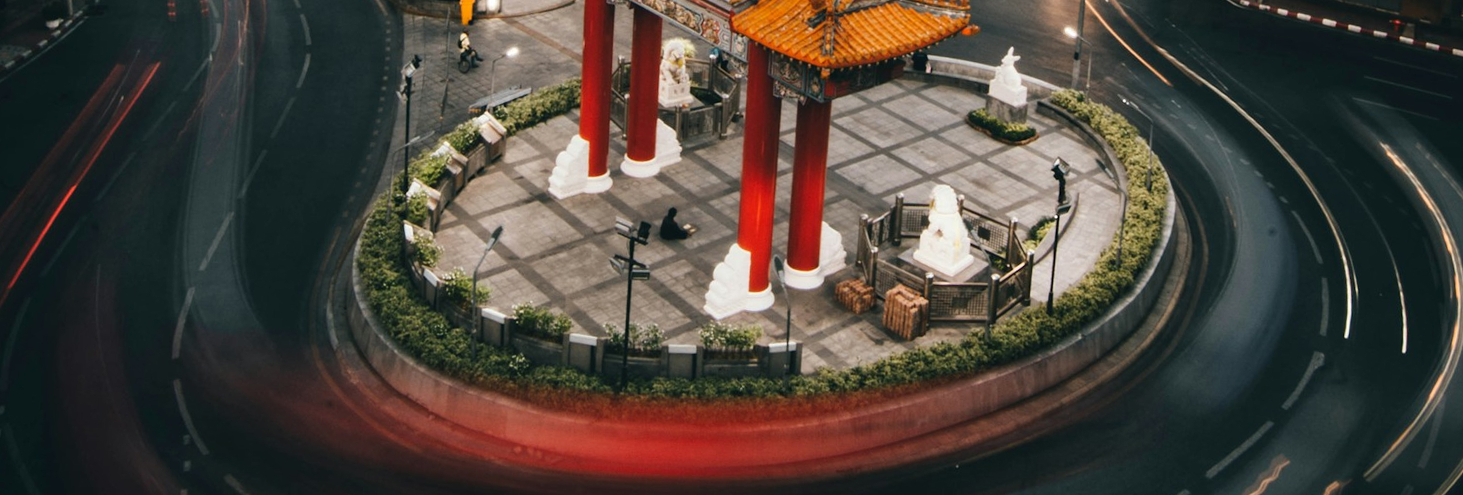 A circular traffic roundabout with a traditional red and orange Chinese pavilion in the center, surrounded by stone lion statues and greenery, with light streaks from passing cars around it.