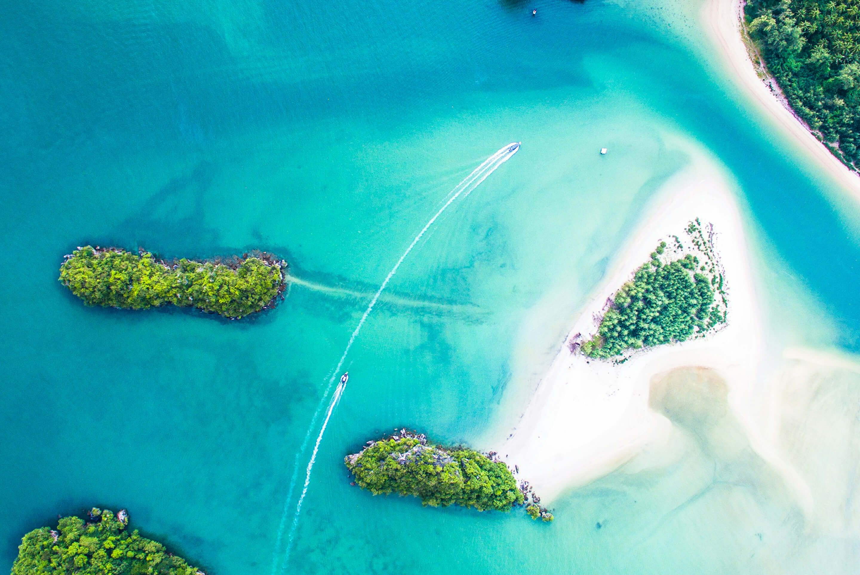 Aerial view of three small green islands surrounded by turquoise water and white sandy beaches with two boats creating trails in the water.