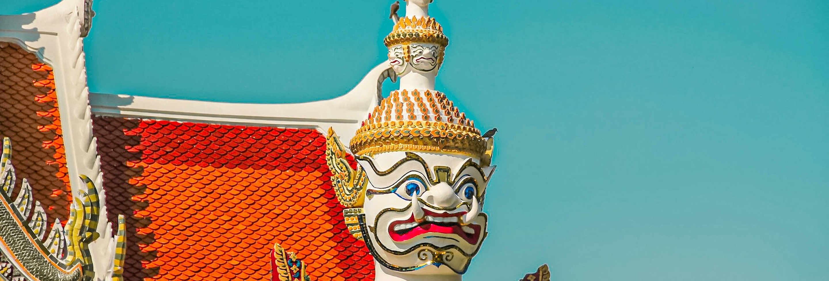 Close-up of an ornate, colorful guardian statue with a white face, blue eyes, and red mouth in front of a red-tiled temple roof and clear blue sky.