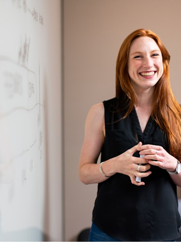 Smiling woman with long red hair standing and holding a whiteboard marker in front of a whiteboard.