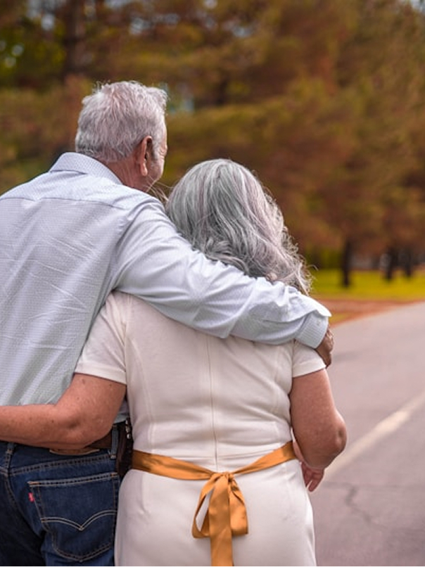 Elderly couple walking outdoors with the man’s arm around the woman amid autumn trees.