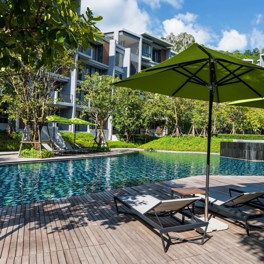 Outdoor swimming pool area with lounge chairs and green umbrellas in front of a modern condo building surrounded by trees.