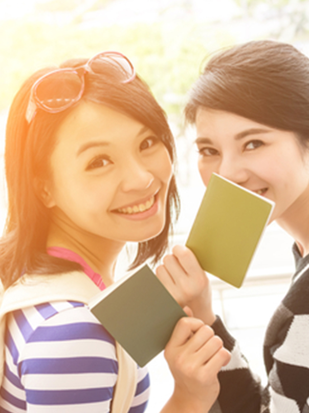 Two smiling young women holding green passports, one wearing striped shirt and sunglasses on her head.