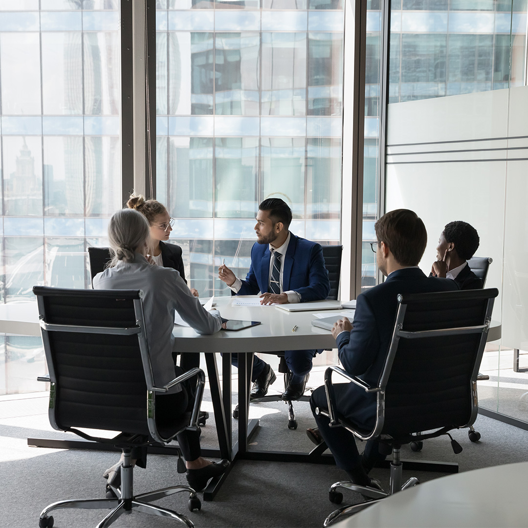 Five professionals in business attire having a meeting around a table in a modern office with large windows.