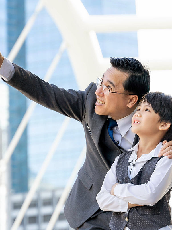 Man in suit and glasses smiling and pointing while standing next to a smiling boy with crossed arms, both looking upwards outdoors.