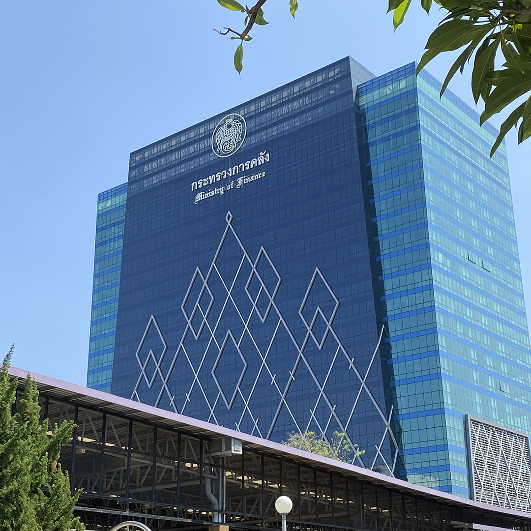 Tall modern glass building with geometric patterns and Ministry of Finance logo and text in Thai and English on facade against clear sky.
